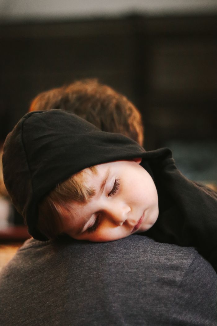 why-choose-us A young child in a hoodie peacefully sleeping on a parent's shoulder indoors.