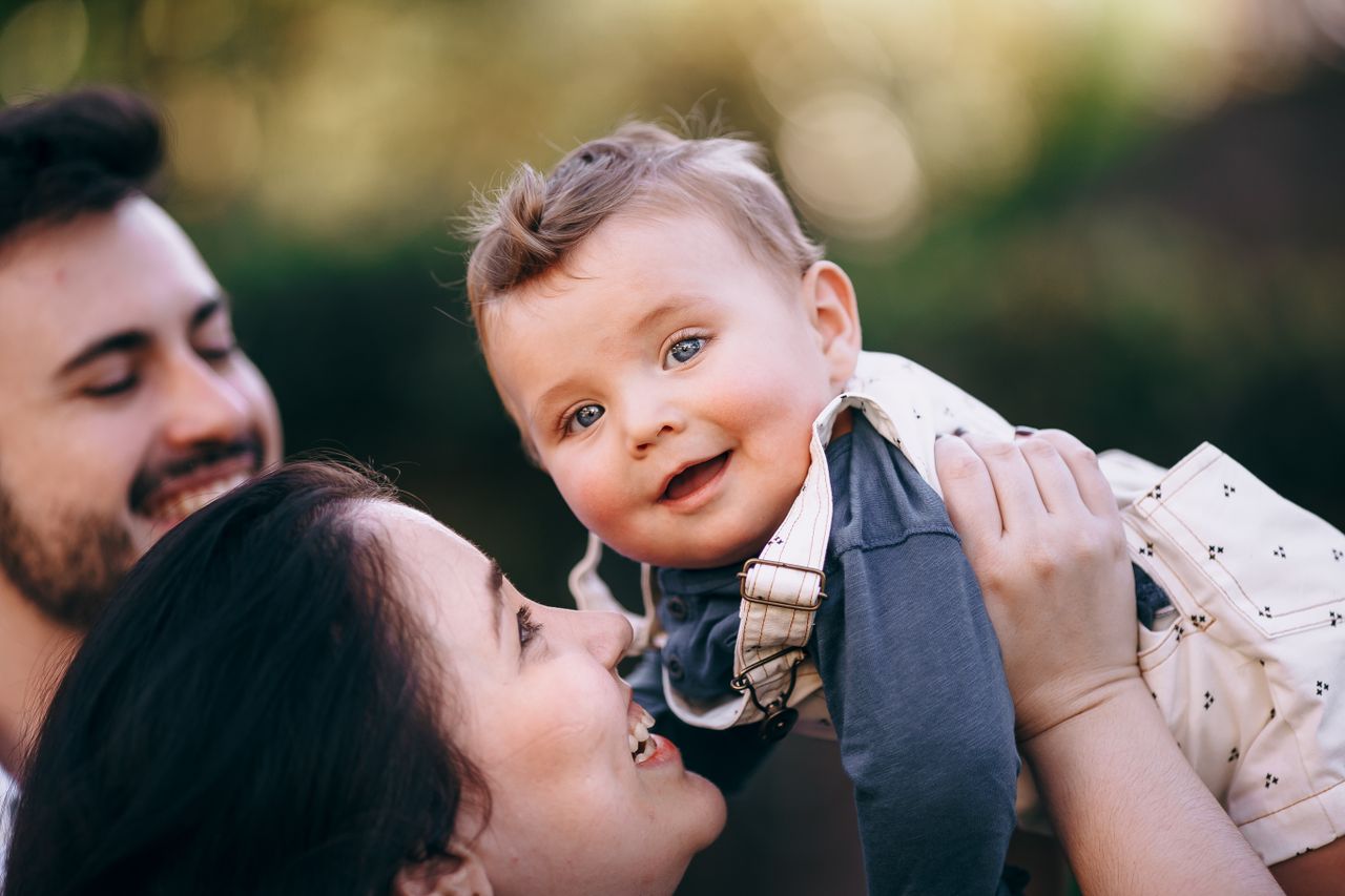 hero-img-01 A joyful family moment with a smiling baby held by parents in a sunny outdoor setting.